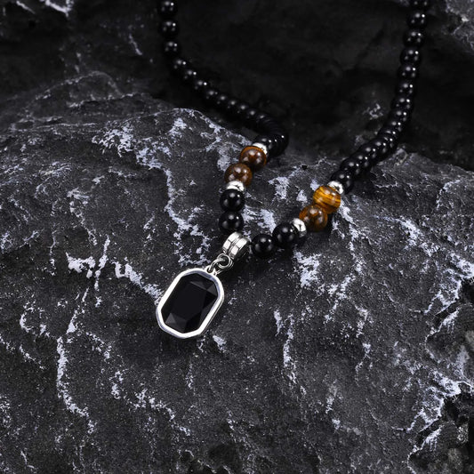 Pendentif rectangulaire noir facetté et perles en pierres naturelles posés sur une roche volcanique sombre.
