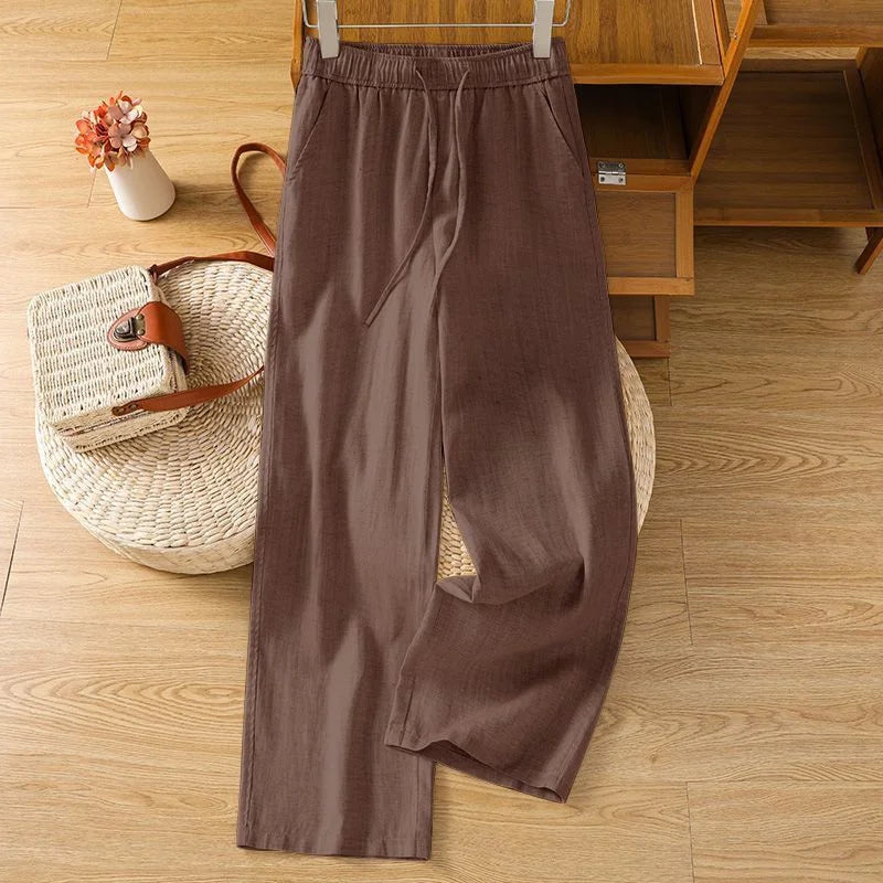 Pantalon large en lin marron sur cintre, posé avec un sac en paille et des fleurs, ambiance estivale.