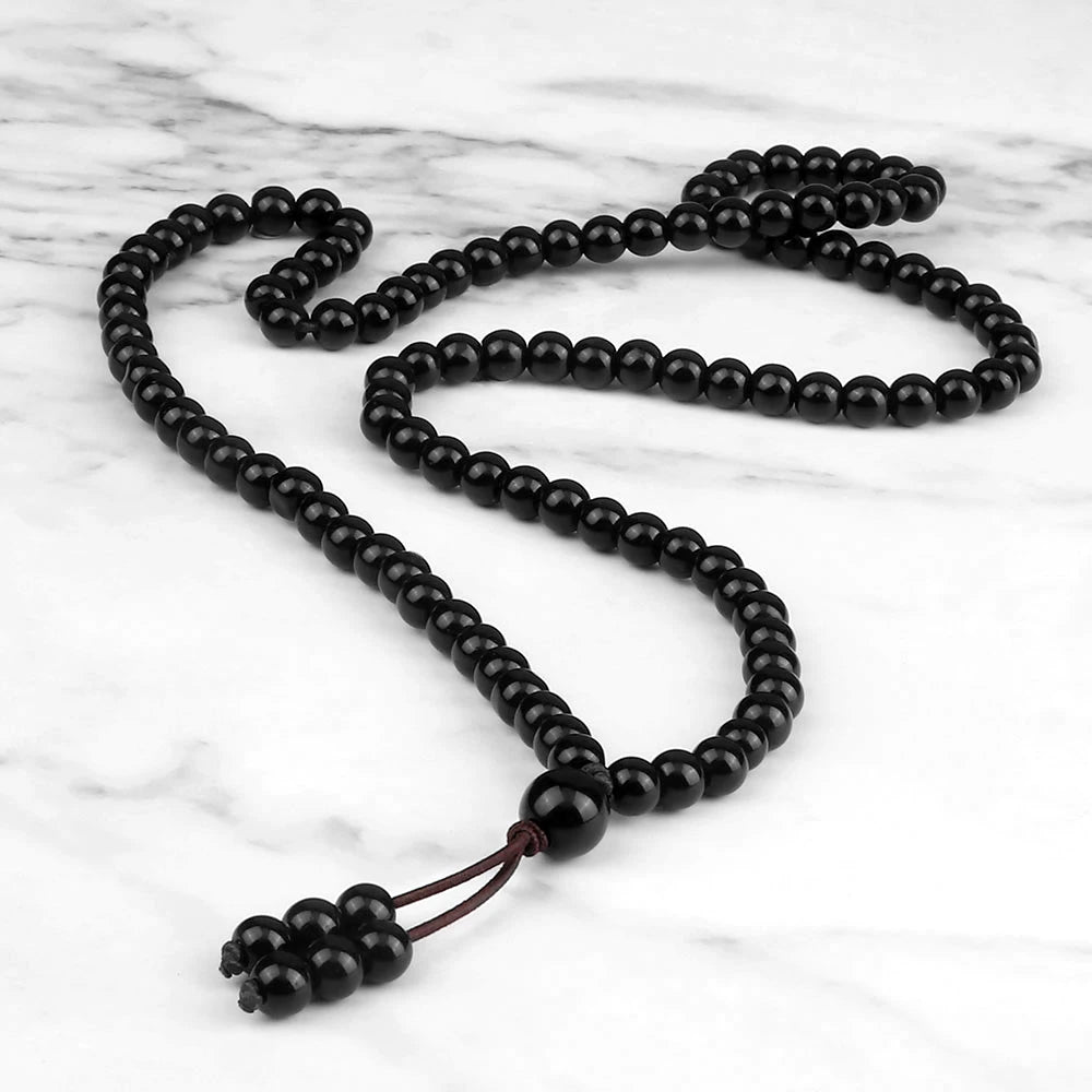 Collier mala 108 perles en pierre noire lisse et brillante, étendu en courbes sur du marbre blanc.