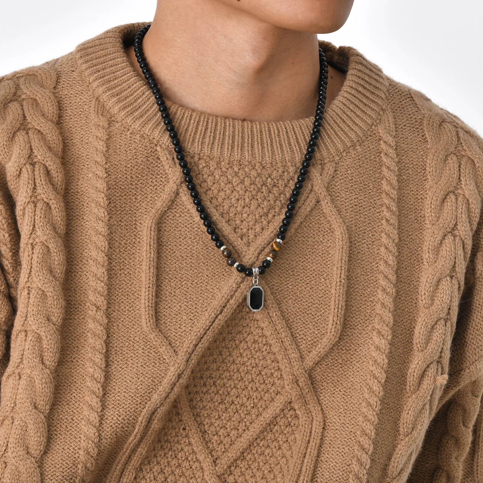 Collier pour homme en perles noires et œil de tigre, porté élégamment sur un pull en maille torsadée marron.